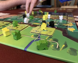 A close-up of a board game featuring colorful wooden pieces in various shapes and colors, placed on a detailed game board. A person’s hand is interacting with the game, indicating active play. The board has a vibrant design with green, yellow, and brown areas.