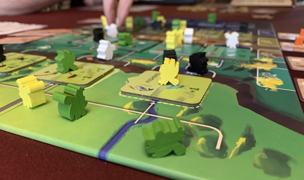A close-up of a board game featuring colorful wooden pieces in various shapes and colors, placed on a detailed game board. A person’s hand is interacting with the game, indicating active play. The board has a vibrant design with green, yellow, and brown areas.