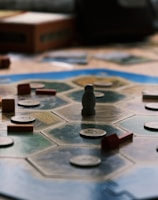 A board game is laid out on a wooden table with hexagonal tiles and various wooden pieces, including houses and roads. Some tiles have numerical tokens on them. There is a box in the background, along with cards and a mobile phone nearby.