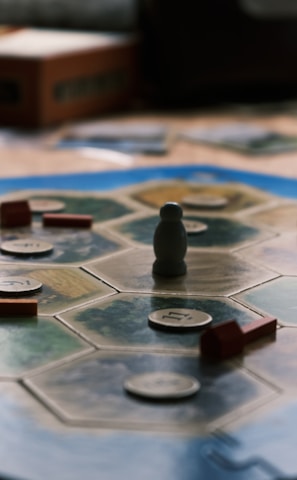 A board game is laid out on a wooden table with hexagonal tiles and various wooden pieces, including houses and roads. Some tiles have numerical tokens on them. There is a box in the background, along with cards and a mobile phone nearby.