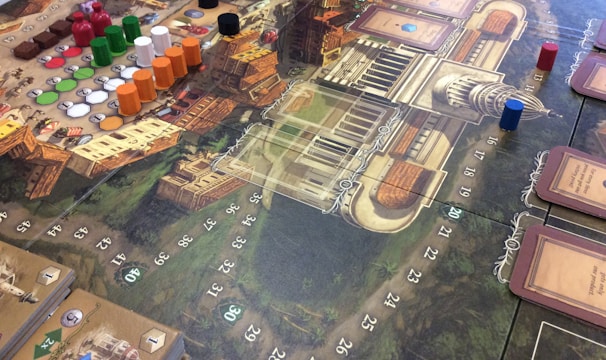 A detailed view of a board game setup with a complex game board featuring a map-like design. Various colored game pieces, including small discs and cylindrical markers in red, green, black, white, and orange, are arranged across the board. There are also decks of cards on the board, each having illustrations and text. The board contains numbered spaces and intricate graphics representing a European city or town. The game appears to be a strategic or historical themed game.