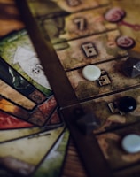 A close-up view of a board game setup, featuring a game board with numbered tiles and various game pieces, including dice and small tokens. Cards in different colors with intricate designs are fanned out beside the board.