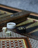 An intricately designed backgammon board with ornate geometric patterns, featuring a pair of dice and playing pieces in close proximity. The board exhibits detailed craftsmanship with inlaid wood work and contrasting colors.