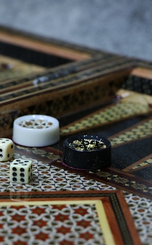 An intricately designed backgammon board with ornate geometric patterns, featuring a pair of dice and playing pieces in close proximity. The board exhibits detailed craftsmanship with inlaid wood work and contrasting colors.