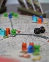 A tabletop game board with colorful wooden animal tokens and two black dice. The board is detailed with winding paths and various game cards propped up in the background.