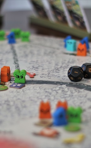 A tabletop game board with colorful wooden animal tokens and two black dice. The board is detailed with winding paths and various game cards propped up in the background.