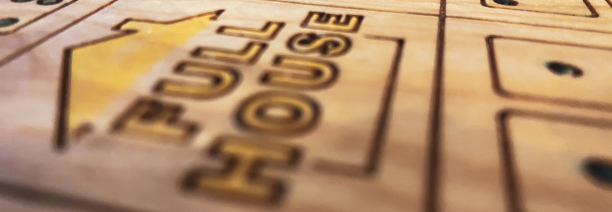 A close-up view of a wooden boardgame surface with engraved patterns, including the words 'FULL HOUSE' and various sections marked with stylized dots resembling dice. The focus is shallow, highlighting the text and some dot patterns while the rest of the board fades softly into the background.