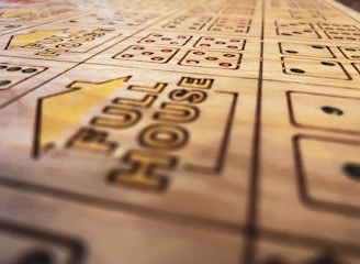 A close-up view of a wooden boardgame surface with engraved patterns, including the words 'FULL HOUSE' and various sections marked with stylized dots resembling dice. The focus is shallow, highlighting the text and some dot patterns while the rest of the board fades softly into the background.