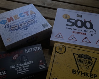 Four different board game boxes placed on a wooden surface. The designs feature colorful and graphic artwork. Each box appears to have distinct themes, represented by unique titles and illustrations.