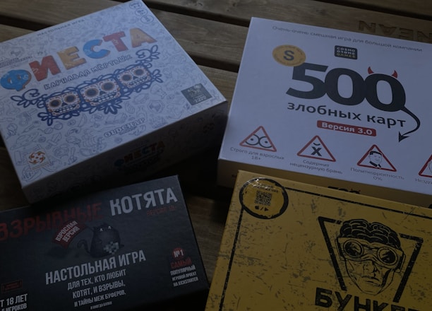Four different board game boxes placed on a wooden surface. The designs feature colorful and graphic artwork. Each box appears to have distinct themes, represented by unique titles and illustrations.